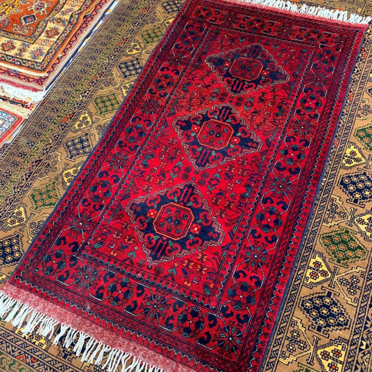 Red patterned rug with white fringe on a collection of other rugs.
